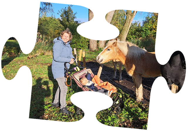 Een jong kind in een roze jasje zit in een rood wagentje, getrokken door een jongen op een gele speelgoedtractor.