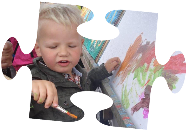 Een jong kind in een roze jasje zit in een rood wagentje, getrokken door een jongen op een gele speelgoedtractor.