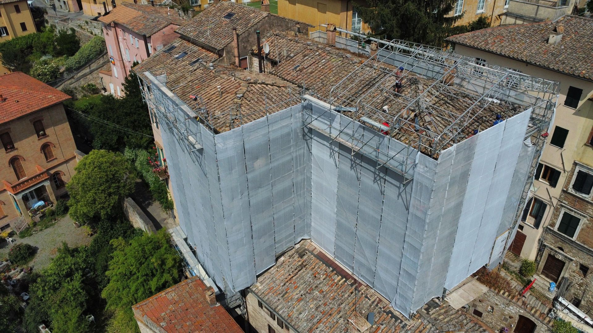 Edificio avvolto da impalcature con tegole a vista, circondato da altri edifici e alberi.