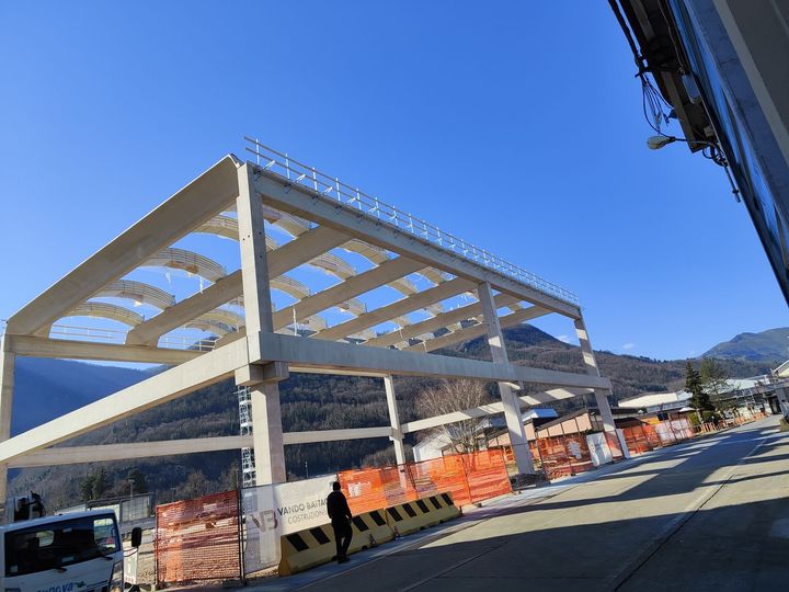 Cantiere con struttura in cemento sotto un cielo azzurro, barriere arancioni, persona in piedi, montagne in lontananza.