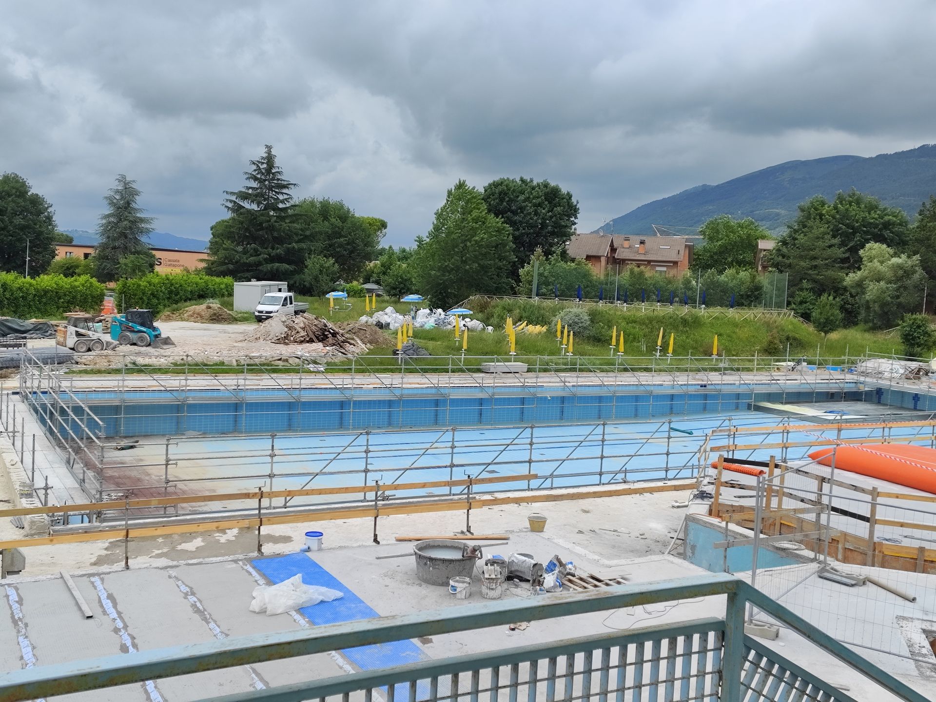 Piscina in costruzione all'aperto con impalcature e interni blu. Alberi verdi e montagne sullo sfondo.