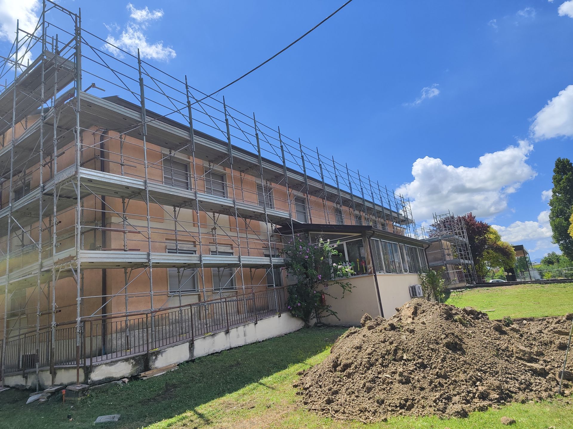 Edificio in ristrutturazione con impalcature, circondato da erba verde e un cumulo di terra. Cielo azzurro con nuvole in alto.