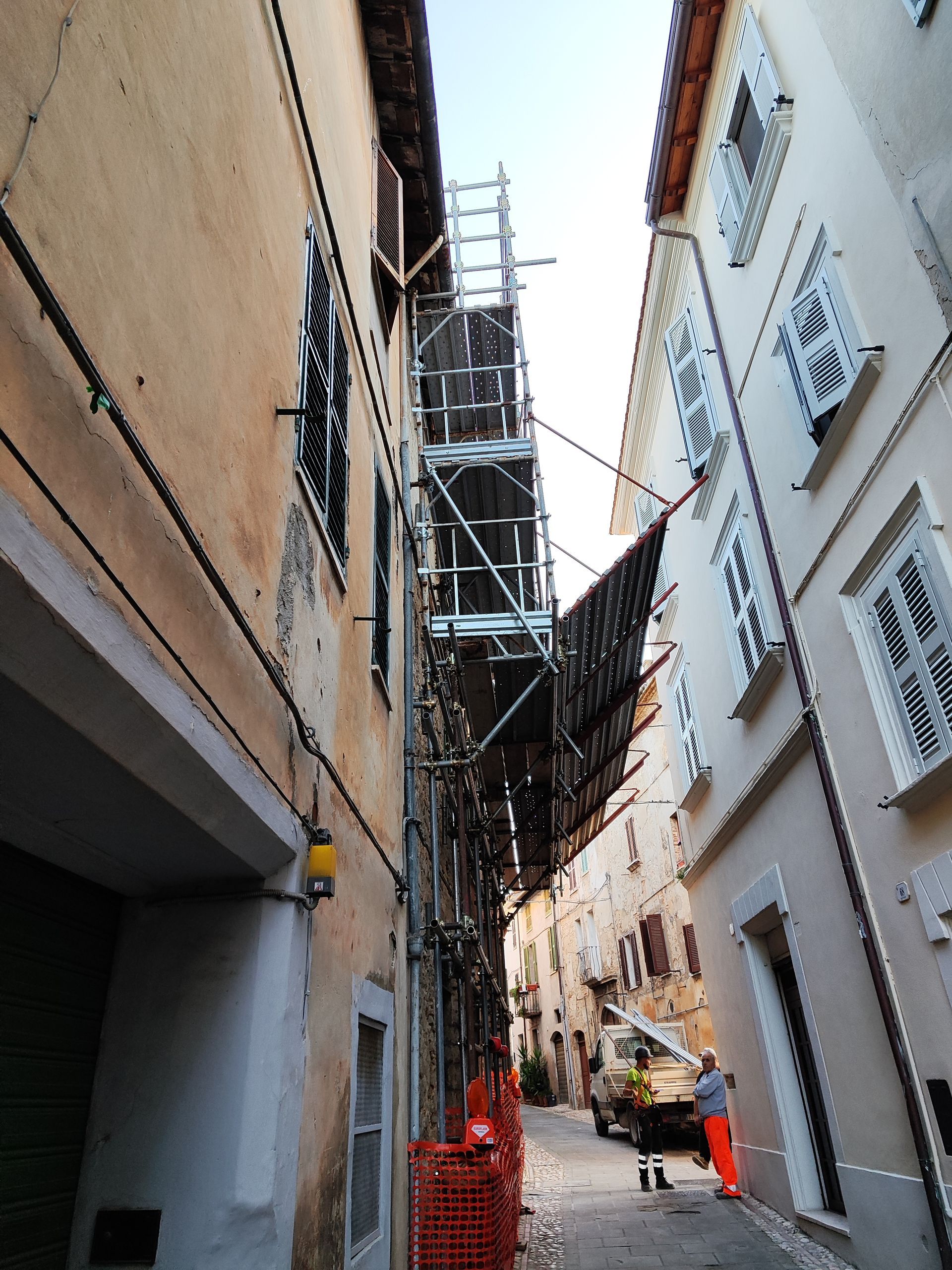 Impalcature tra vecchi edifici in una strada stretta; lavori in corso. Persona in arancione visibile.