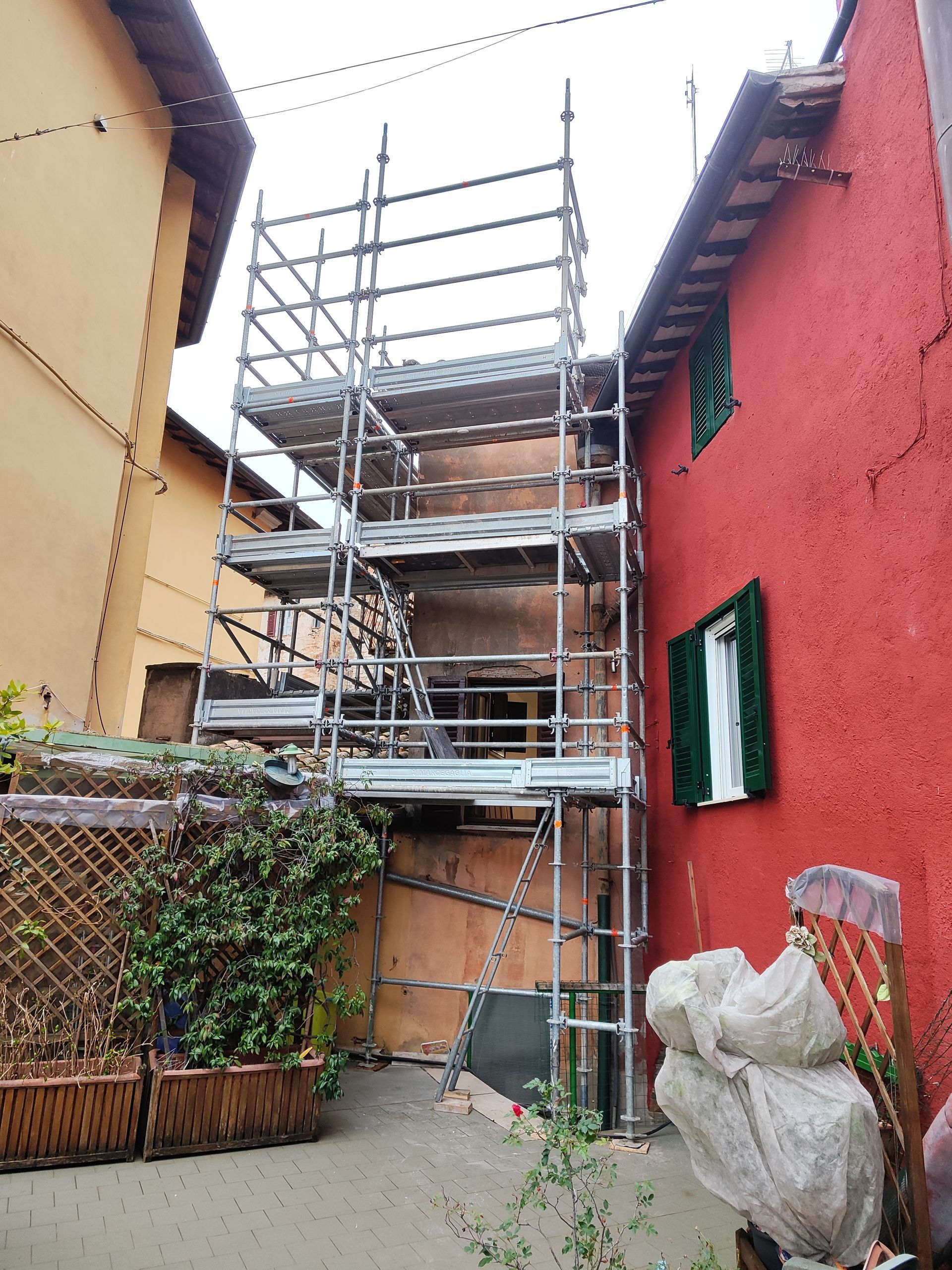 Impalcatura all'esterno di un edificio con pareti rosse e gialle, una persiana verde e una fioriera in legno.