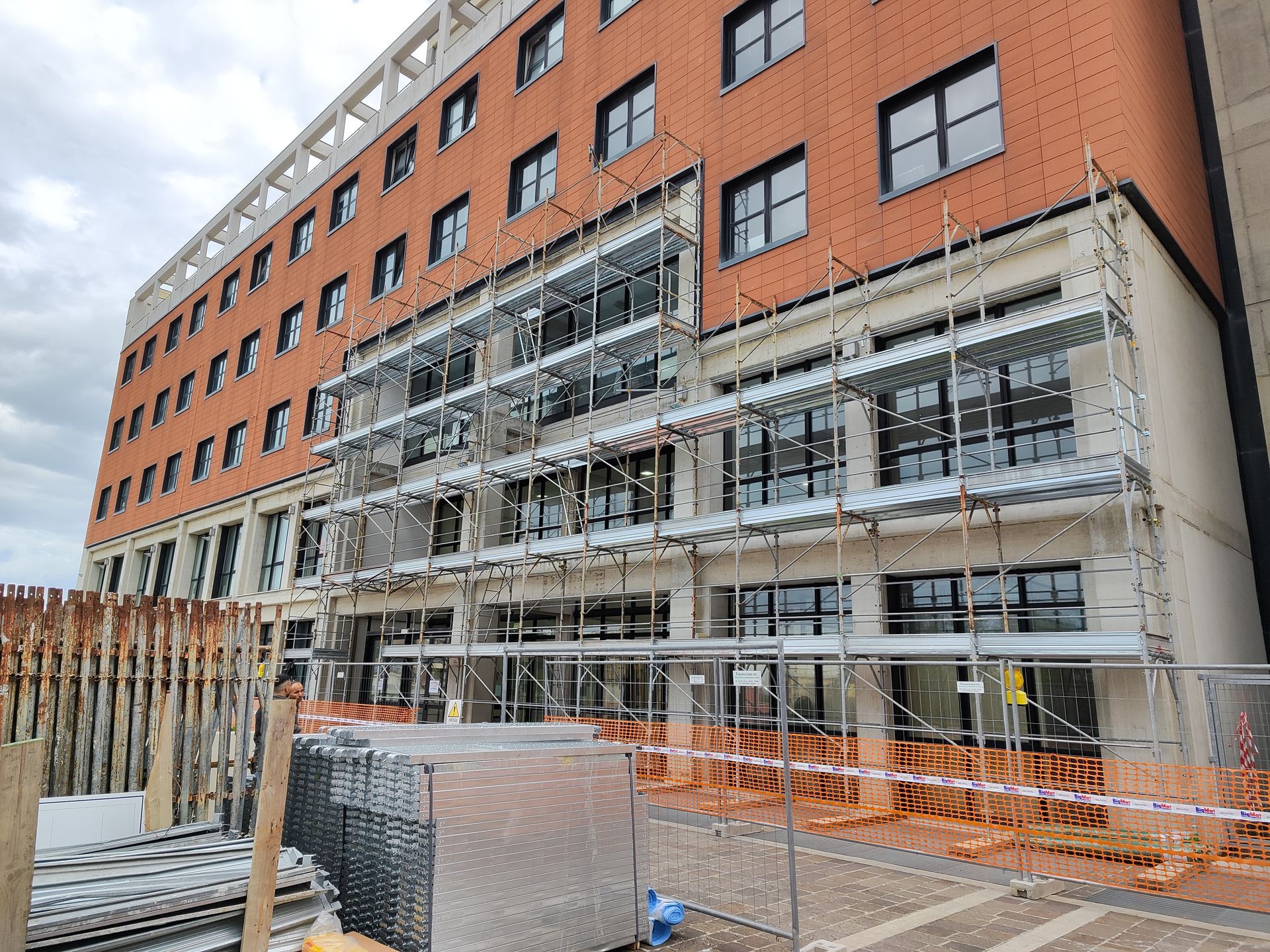 Edificio in costruzione con impalcature. Facciata in mattoni, finestre e recinzione di sicurezza visibili.