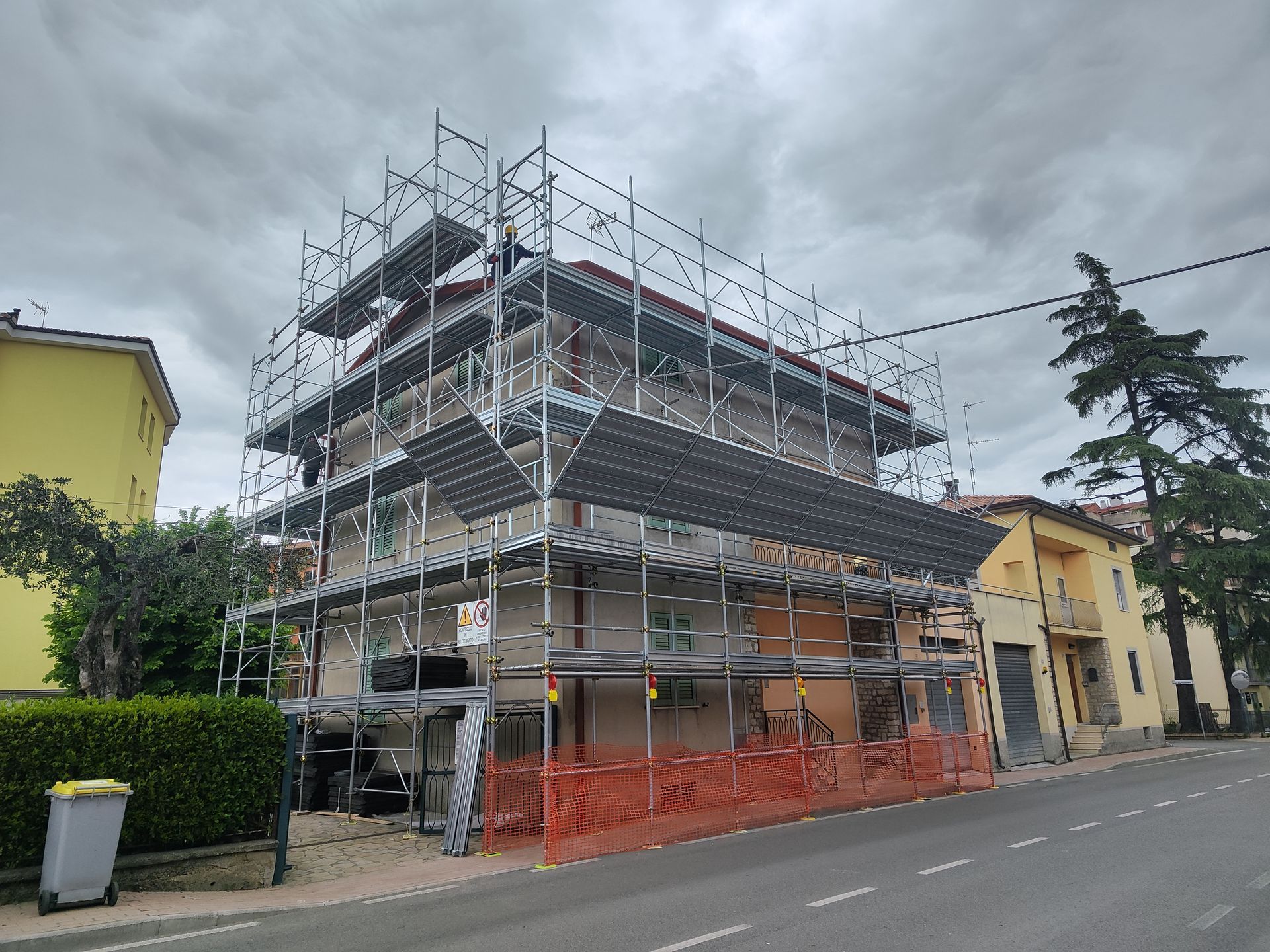 Edificio di tre piani in costruzione con impalcature. Recinzione di sicurezza arancione a terra. Cielo grigio e nuvoloso.