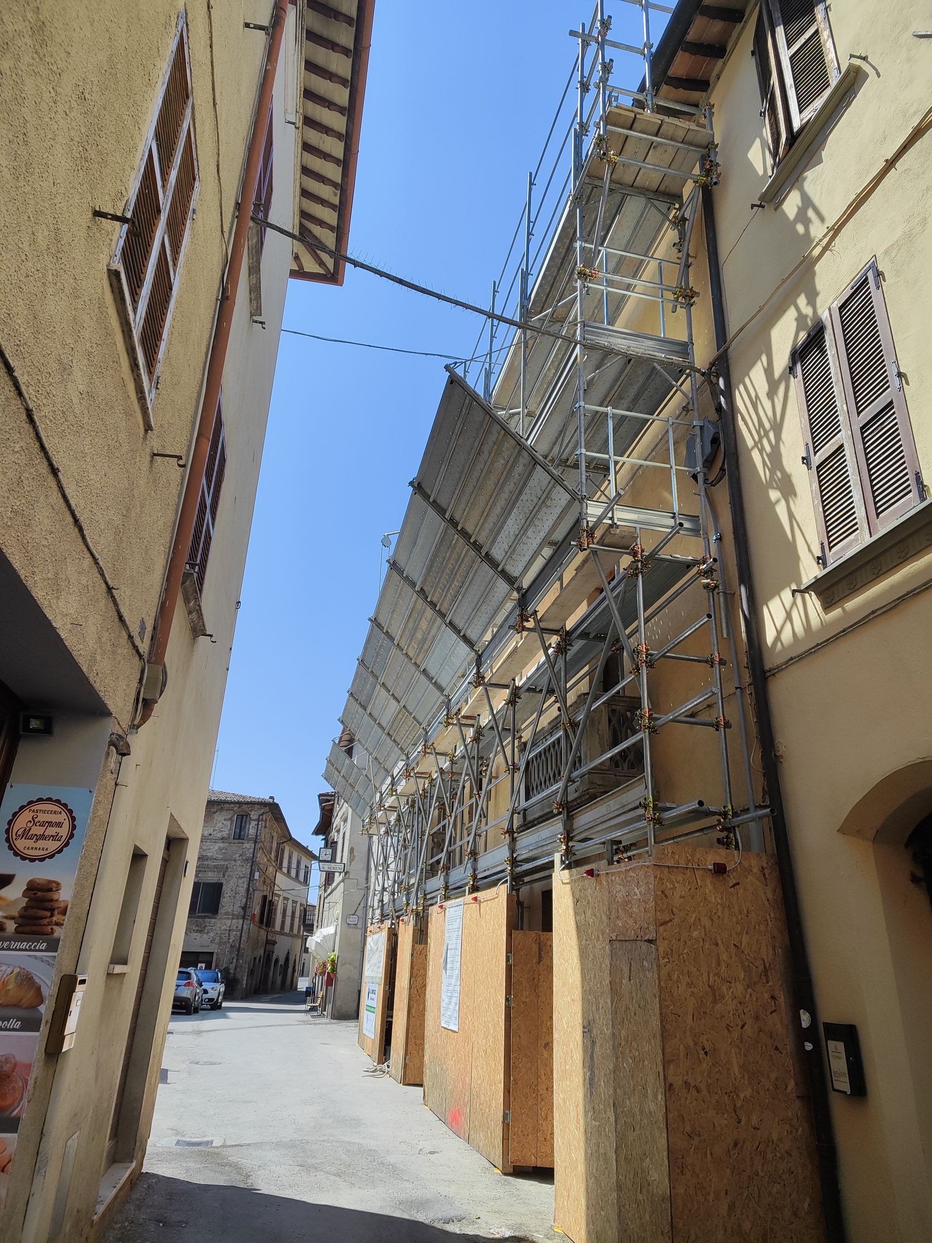 Strada stretta con impalcature sul lato di un edificio, strutture alte e persiane marroni.