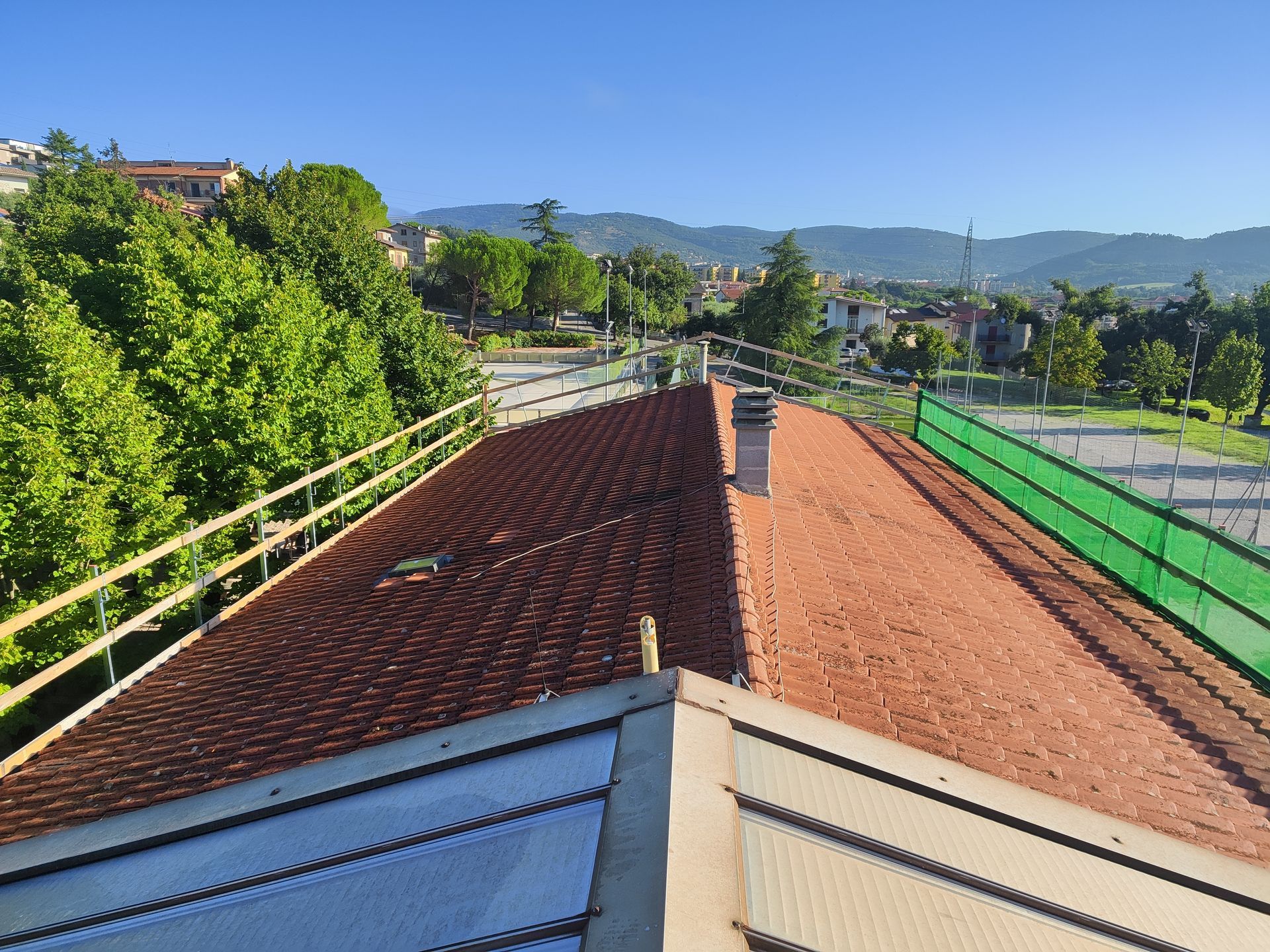 Vista di un tetto in tegole di terracotta con ringhiere di sicurezza, che si affaccia su un parco e una zona residenziale sotto un cielo azzurro.