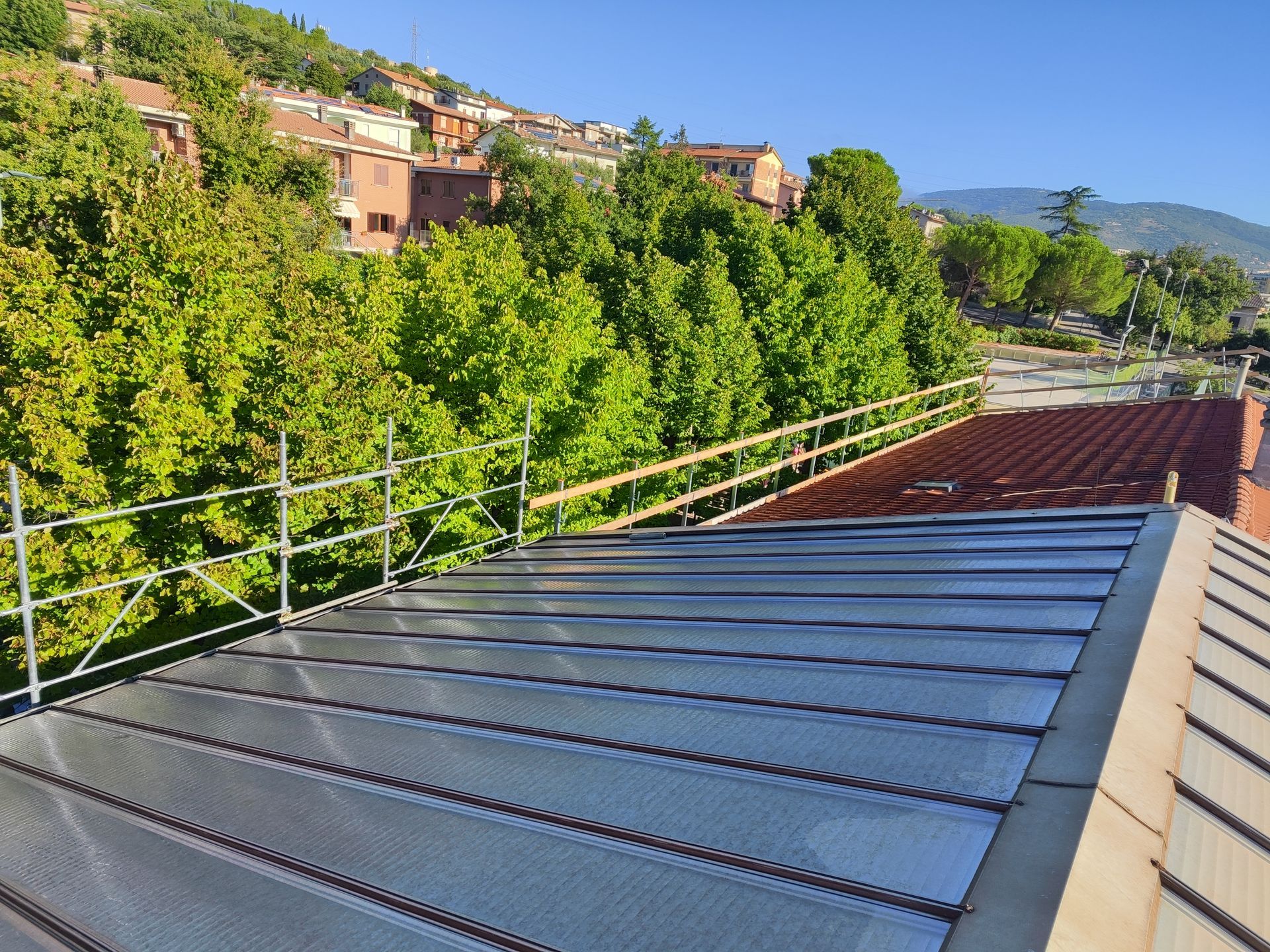 Tetto con pannelli metallici e ringhiere di sicurezza, alberi e case sullo sfondo in una giornata di sole.