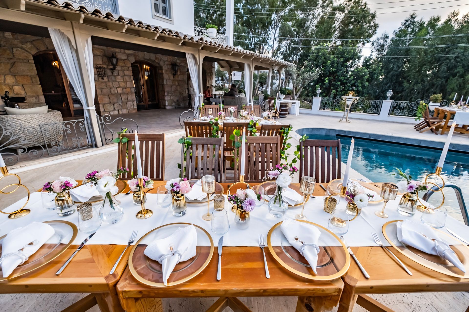 A long wooden table is set for a wedding reception in front of a swimming pool.
