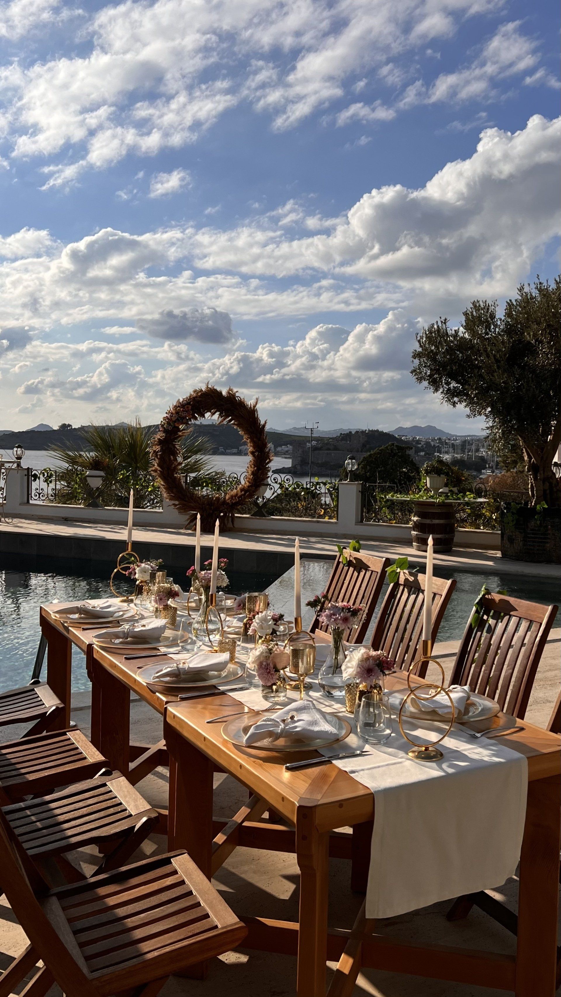A long wooden table is set for a dinner party in front of a swimming pool.
