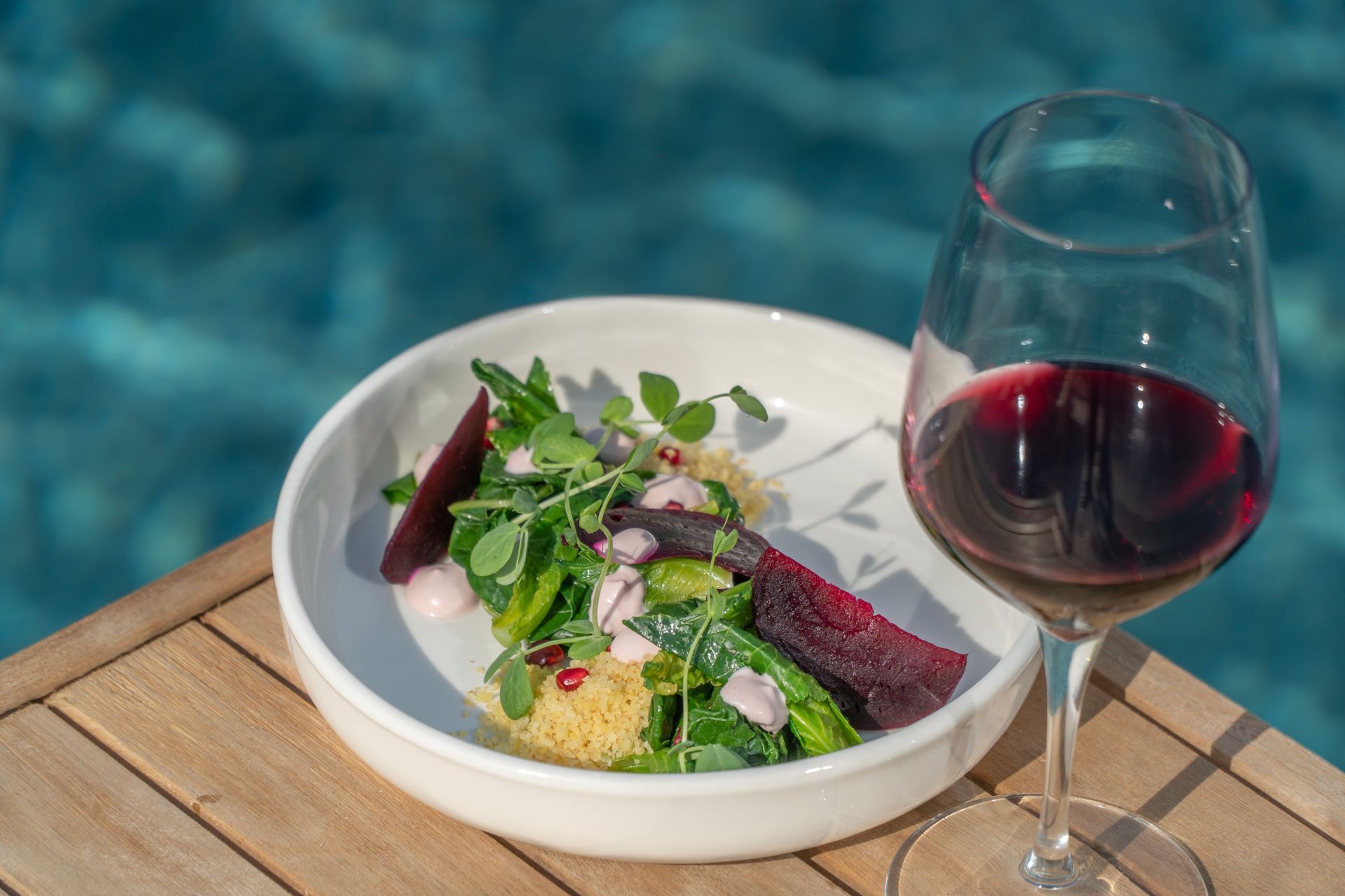 A plate of food and a glass of wine are on a table next to a pool.