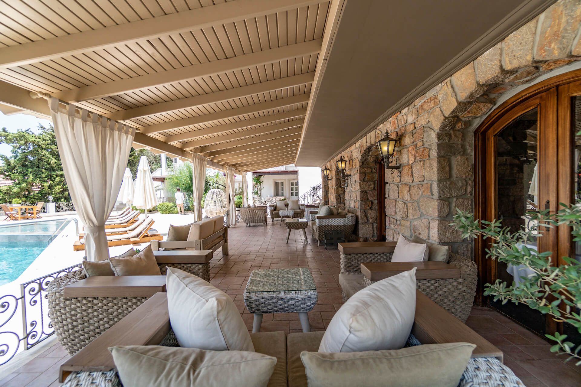 A patio with chairs , tables and curtains overlooking a swimming pool.