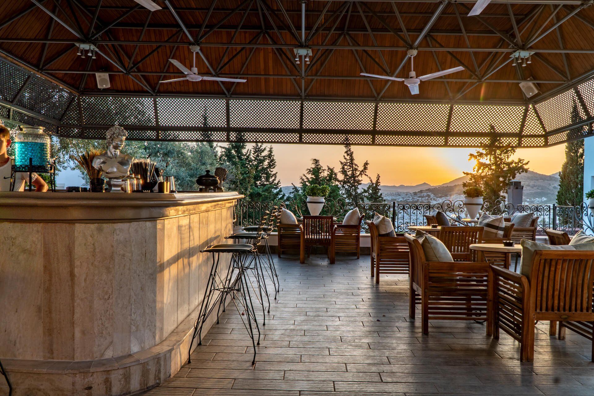 A restaurant with a bar , chairs , tables and a sunset in the background.