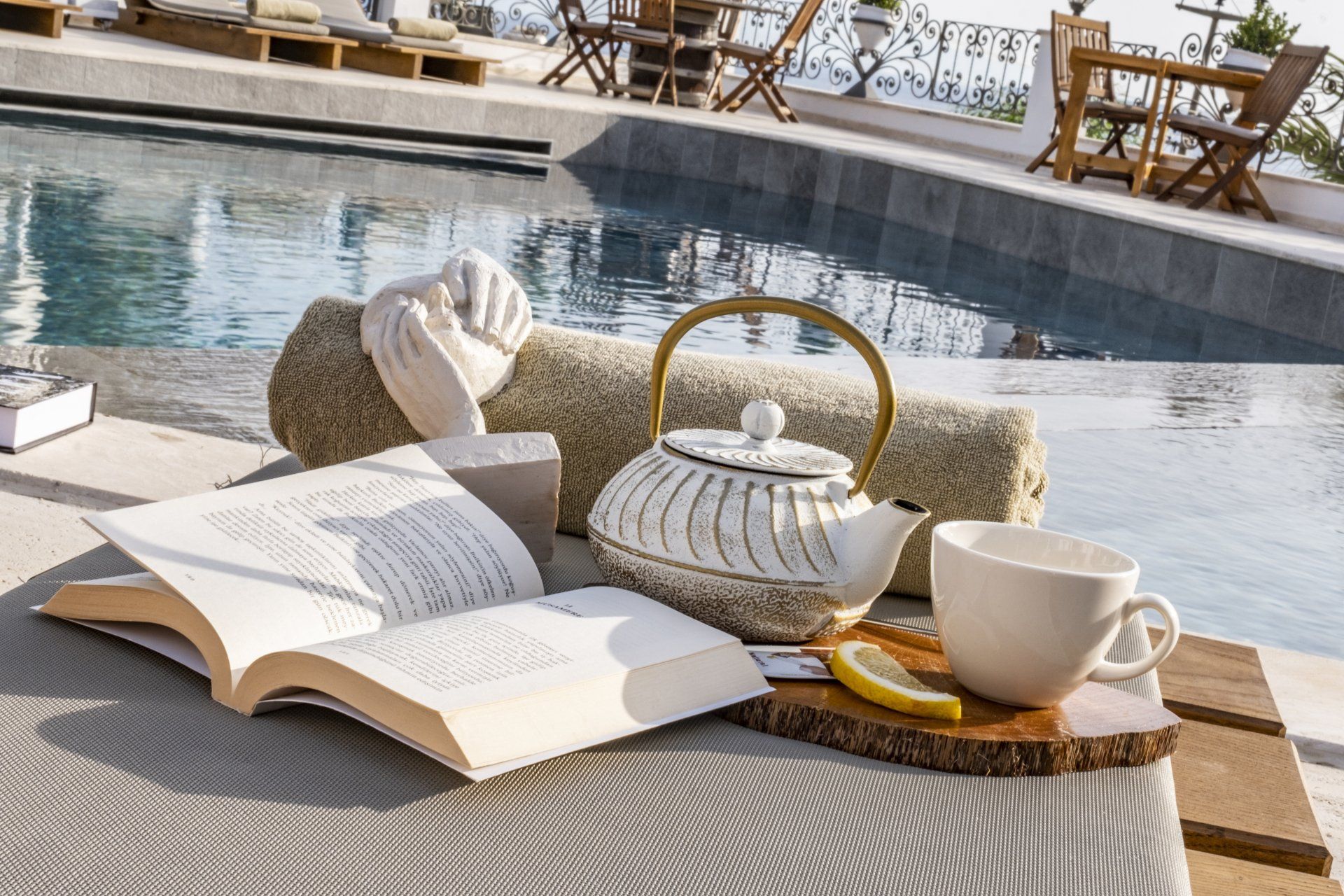 A book , teapot , cup and lemon are on a table next to a pool.