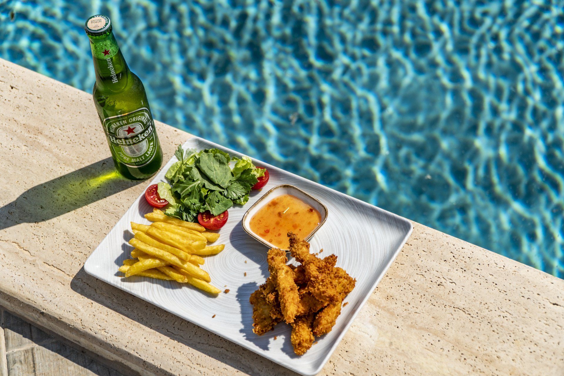 A plate of food and a bottle of beer are sitting next to a swimming pool.