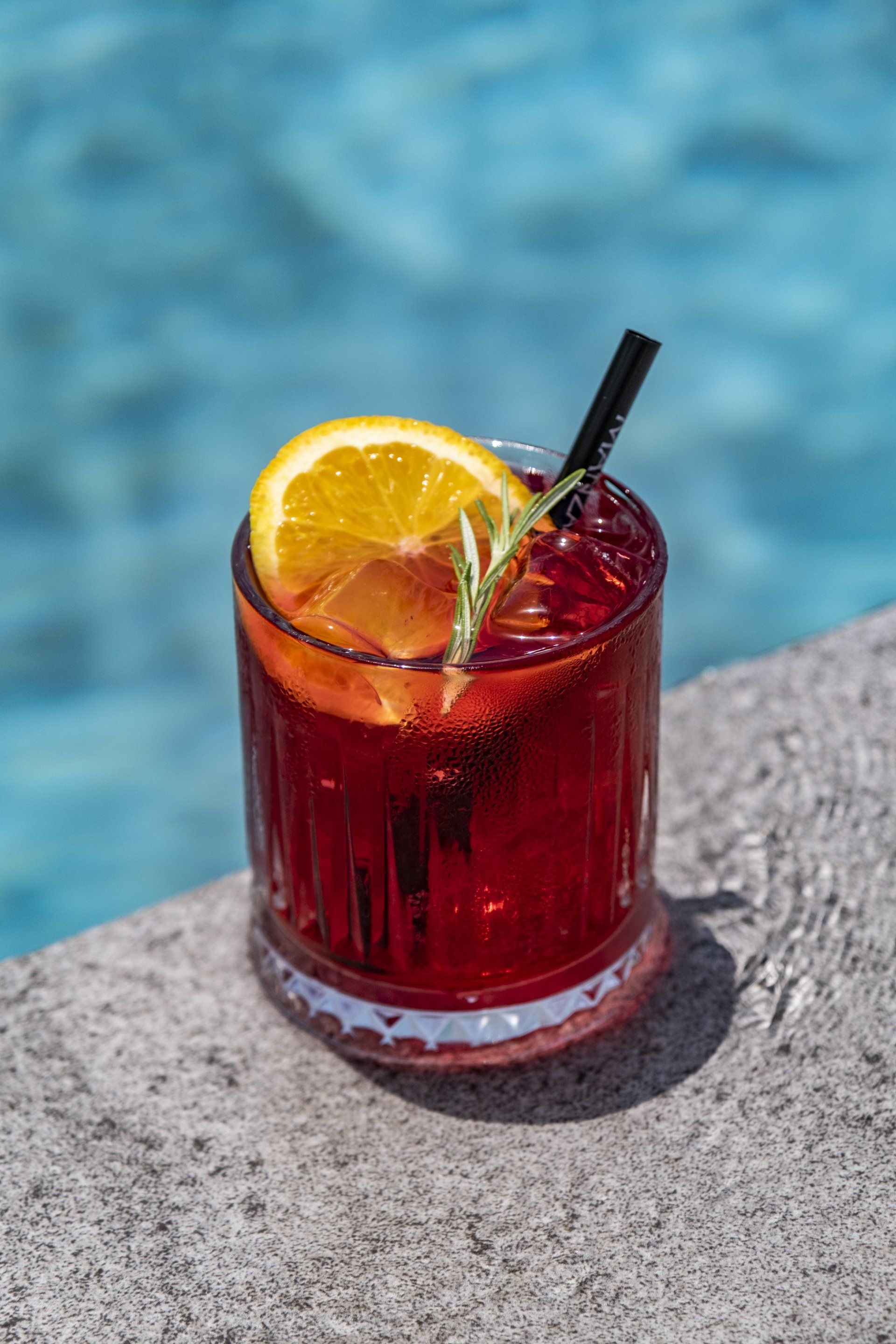 A red cocktail with a straw and a slice of orange is sitting on the edge of a swimming pool.