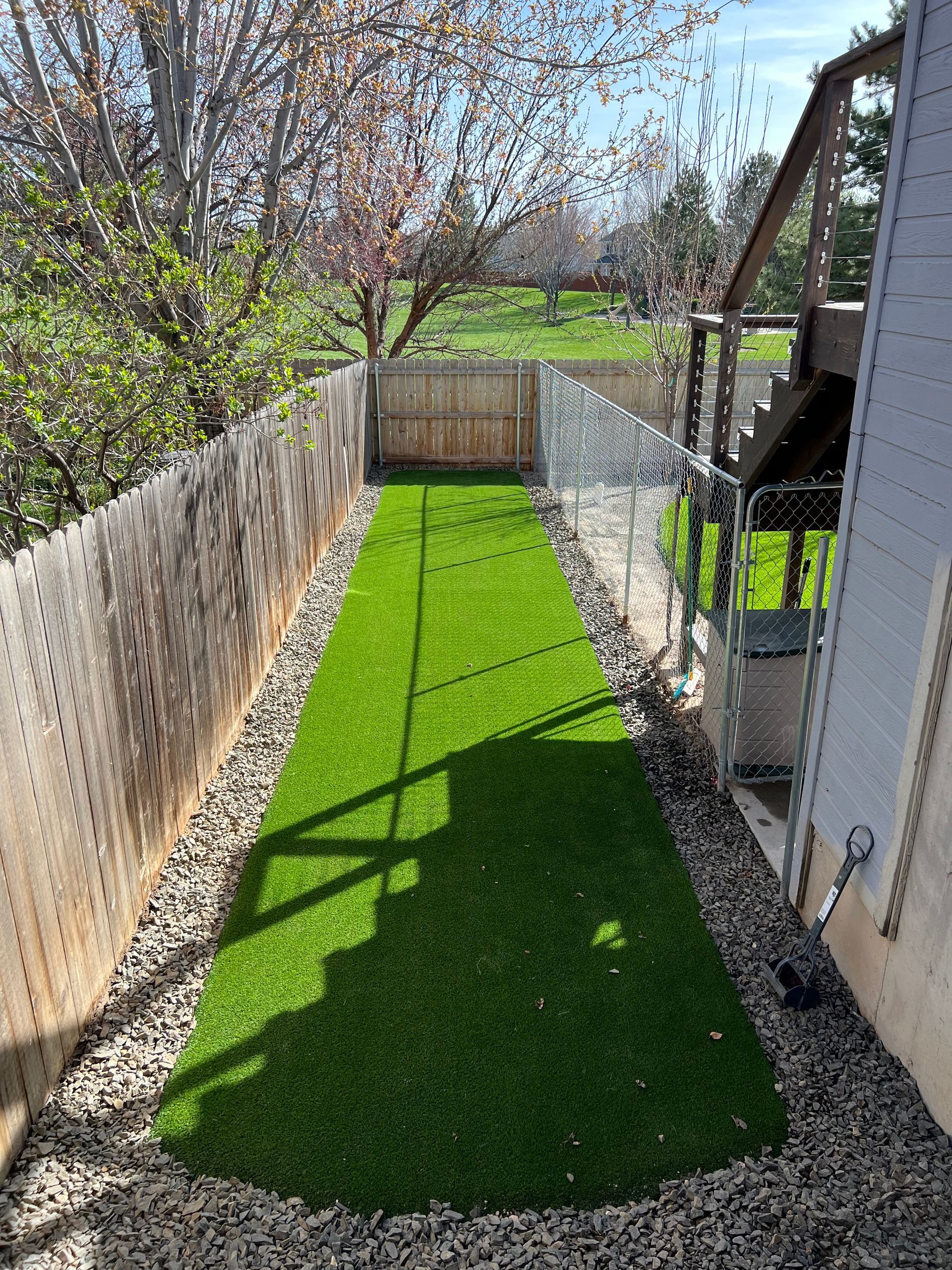 Narrow backyard with artificial turf, bordered by rocks and fences. Sunny day.