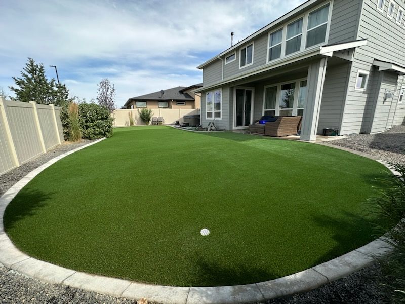 Backyard with artificial turf putting green bordered by concrete, near a two-story house.