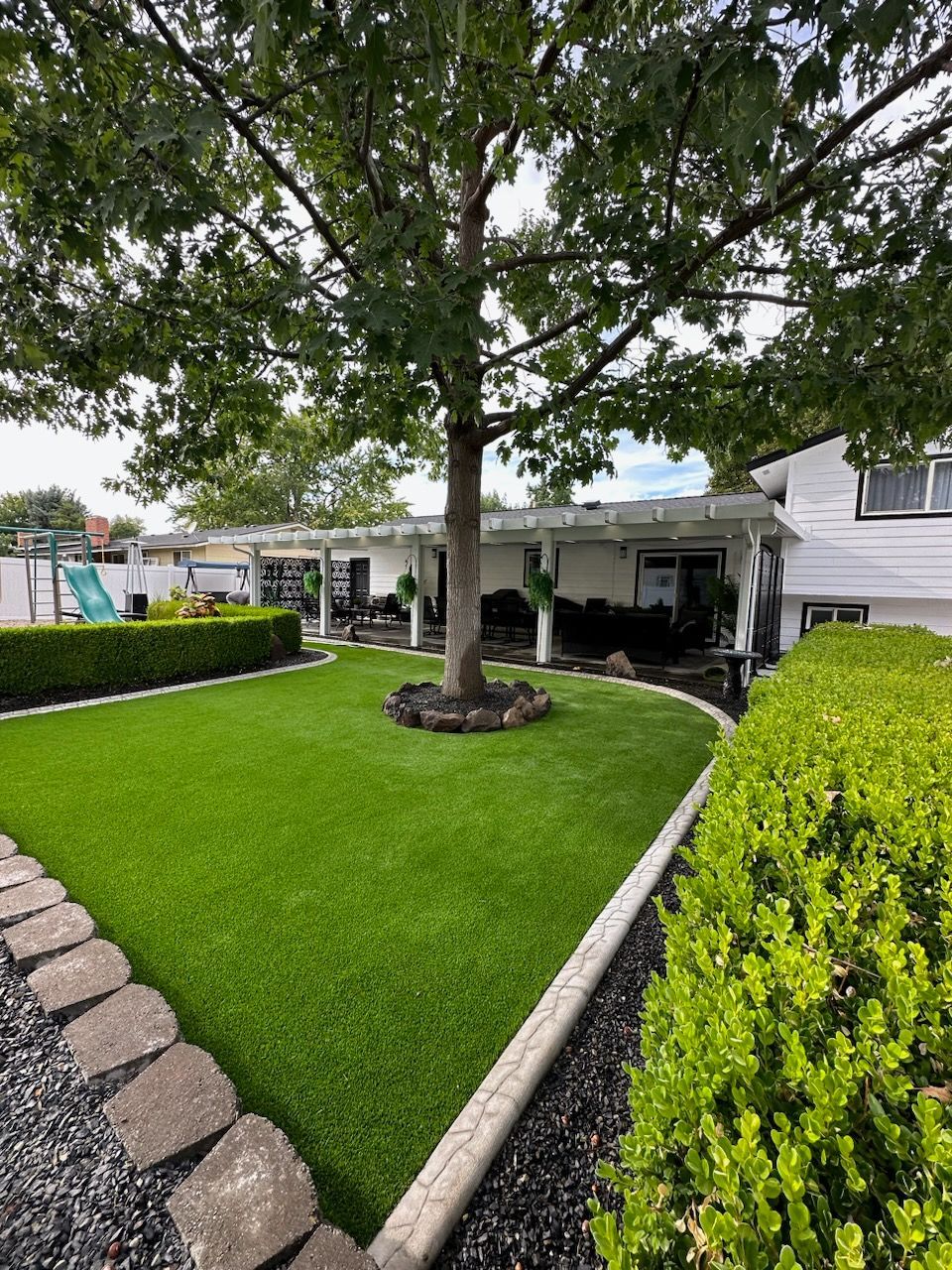 Lush green lawn with tree, bordered by stone and bushes, near a white house with a pergola.