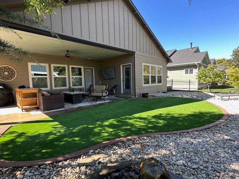 Backyard patio with artificial turf and stone border, beside a light-colored house under a blue sky.