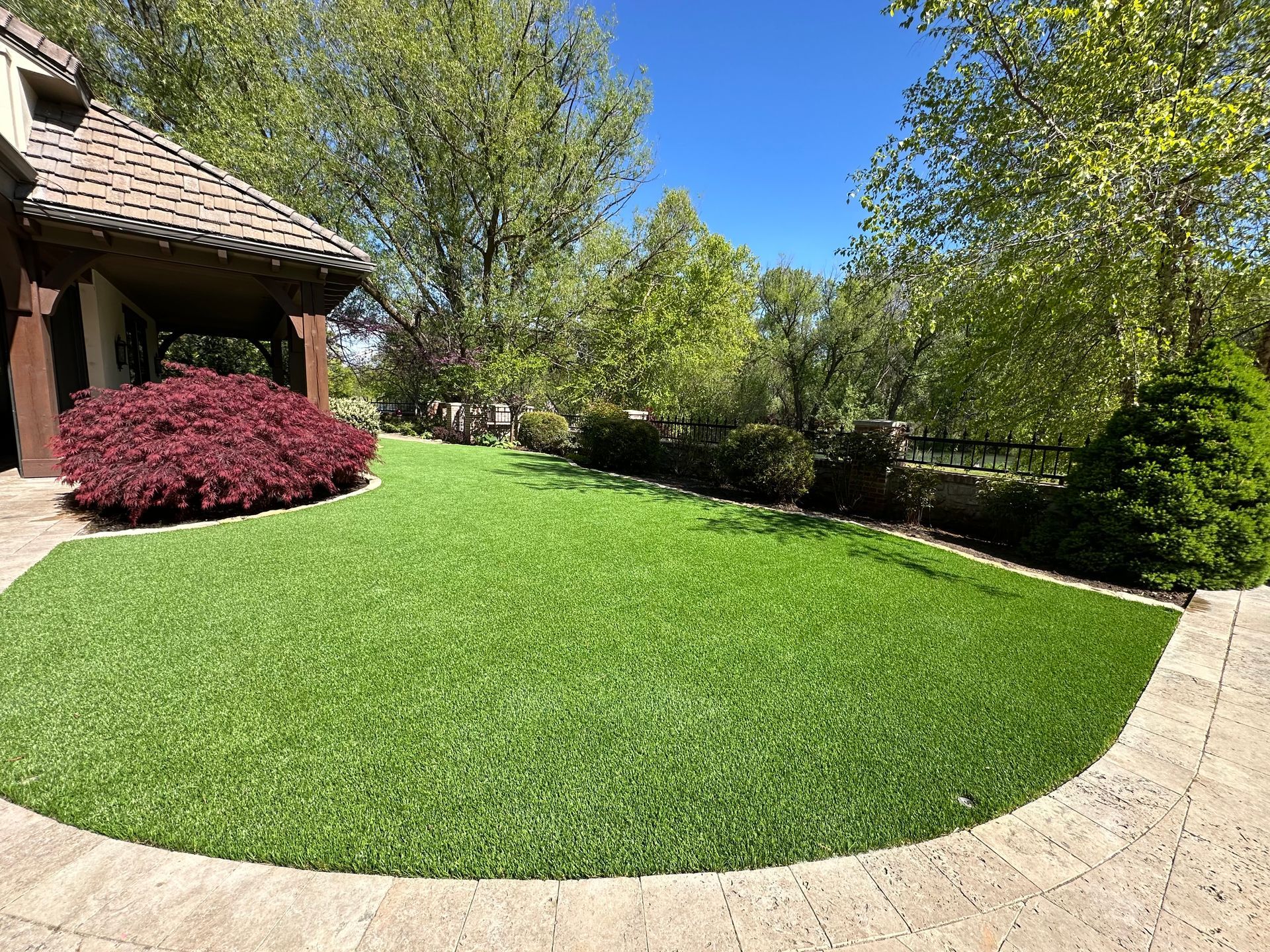 Lush green lawn bordered by stone path, trees, and house with brown roof and red bush under blue sky.