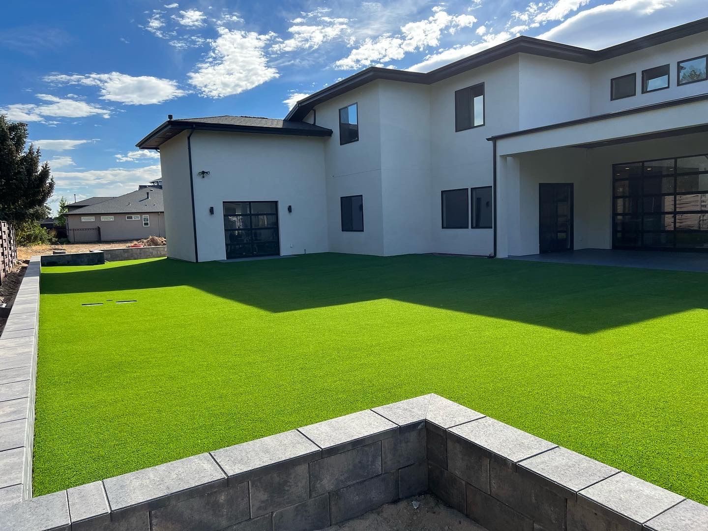 Backyard with bright green artificial grass, modern white house, blue sky with clouds.