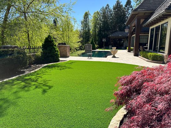 Lush green artificial lawn in a backyard, bordered by a patio, trees, and a house with a red Japanese maple in the foreground.