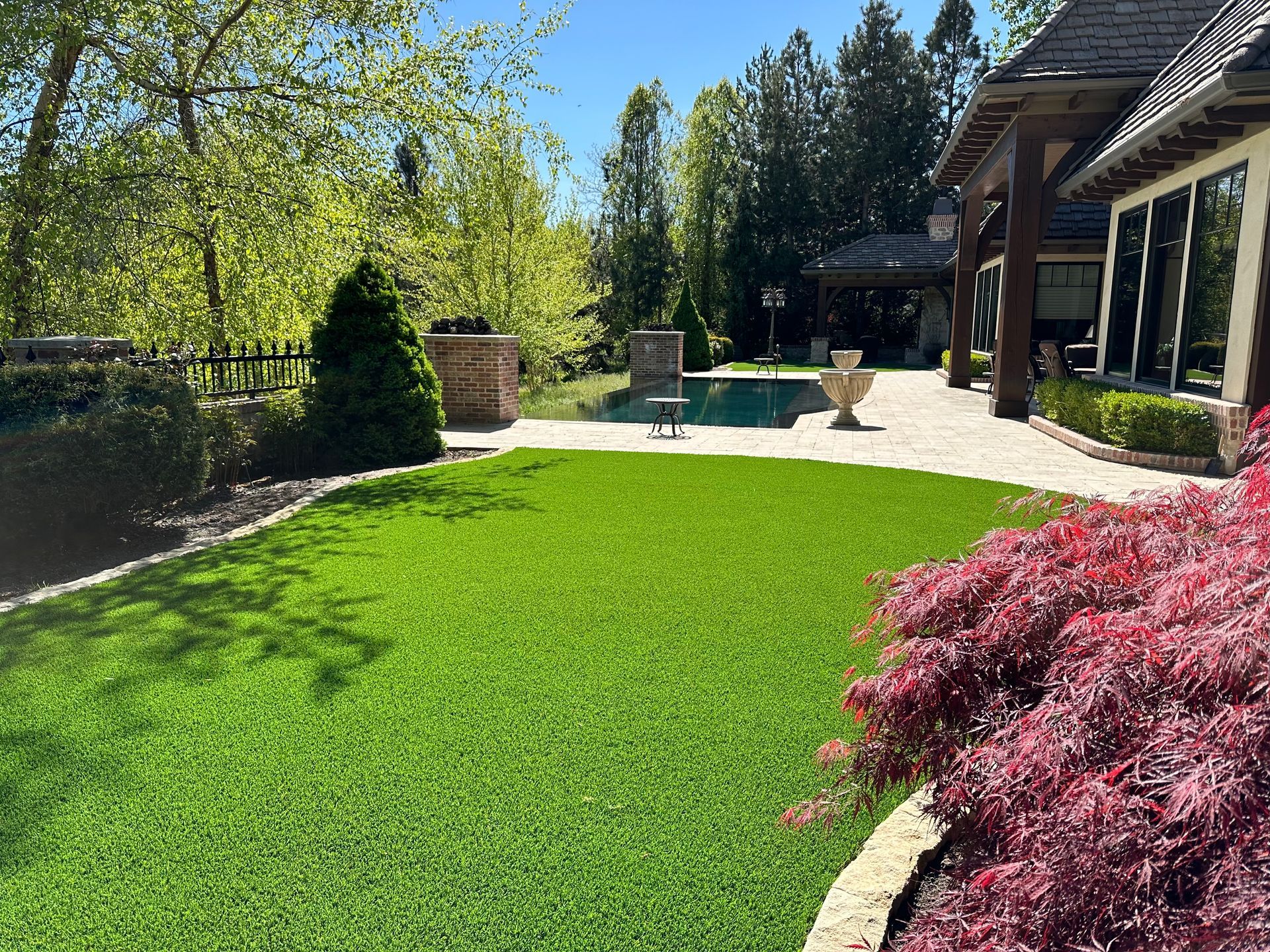 Lush green lawn beside a house with a red-leafed tree in the foreground. Brick patio and trees in the background. Sunny day.