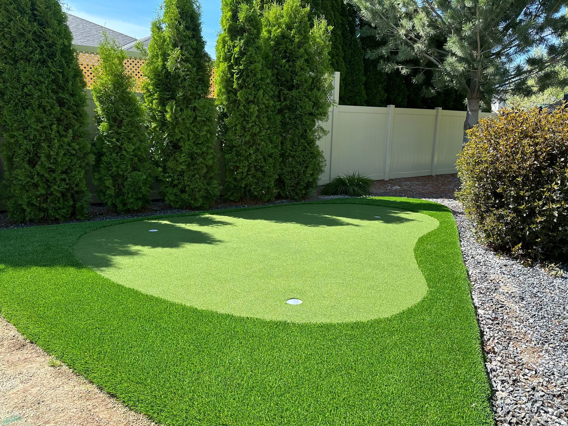 Small putting green surrounded by evergreen trees, with a stone border and a beige fence.