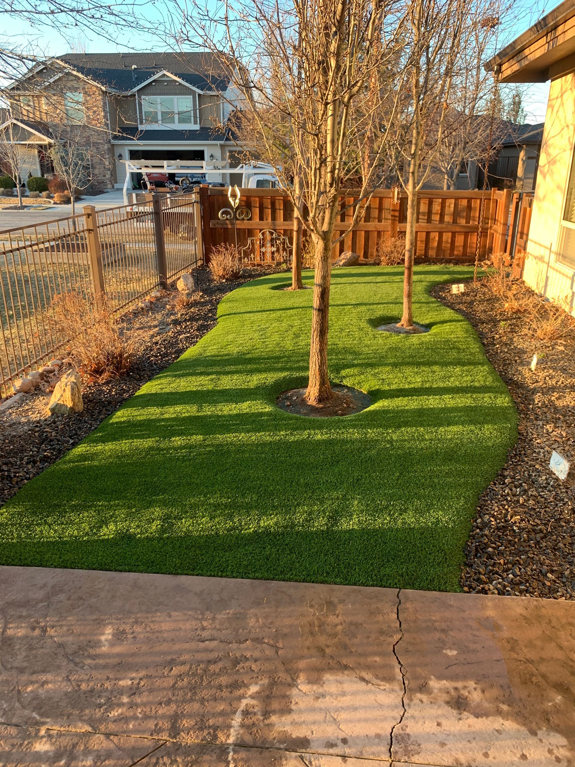 Green artificial turf with trees, brown mulch, and wooden fence in a front yard.