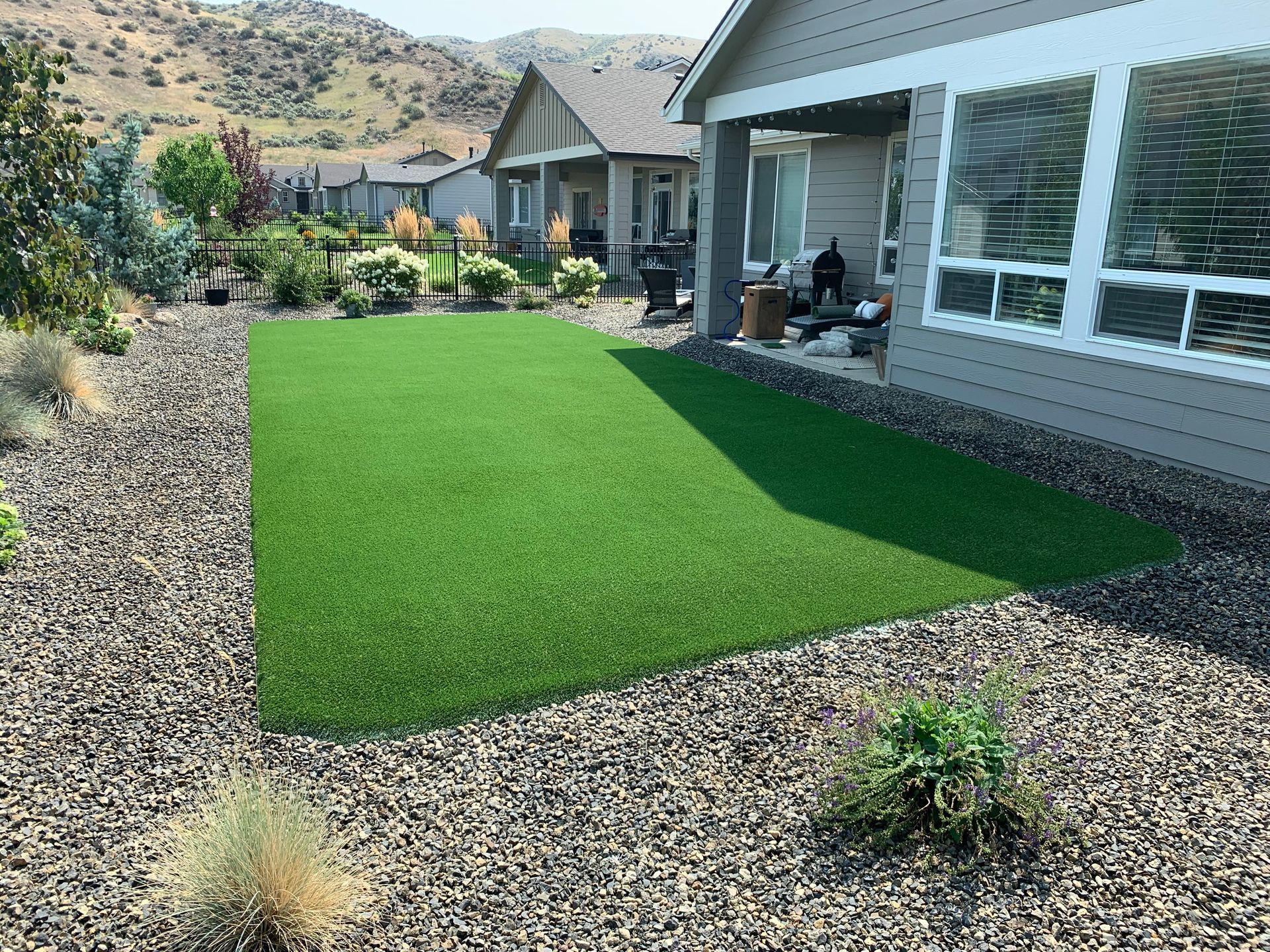 Backyard with artificial green grass, surrounded by gravel and plants, adjacent to a light gray house.