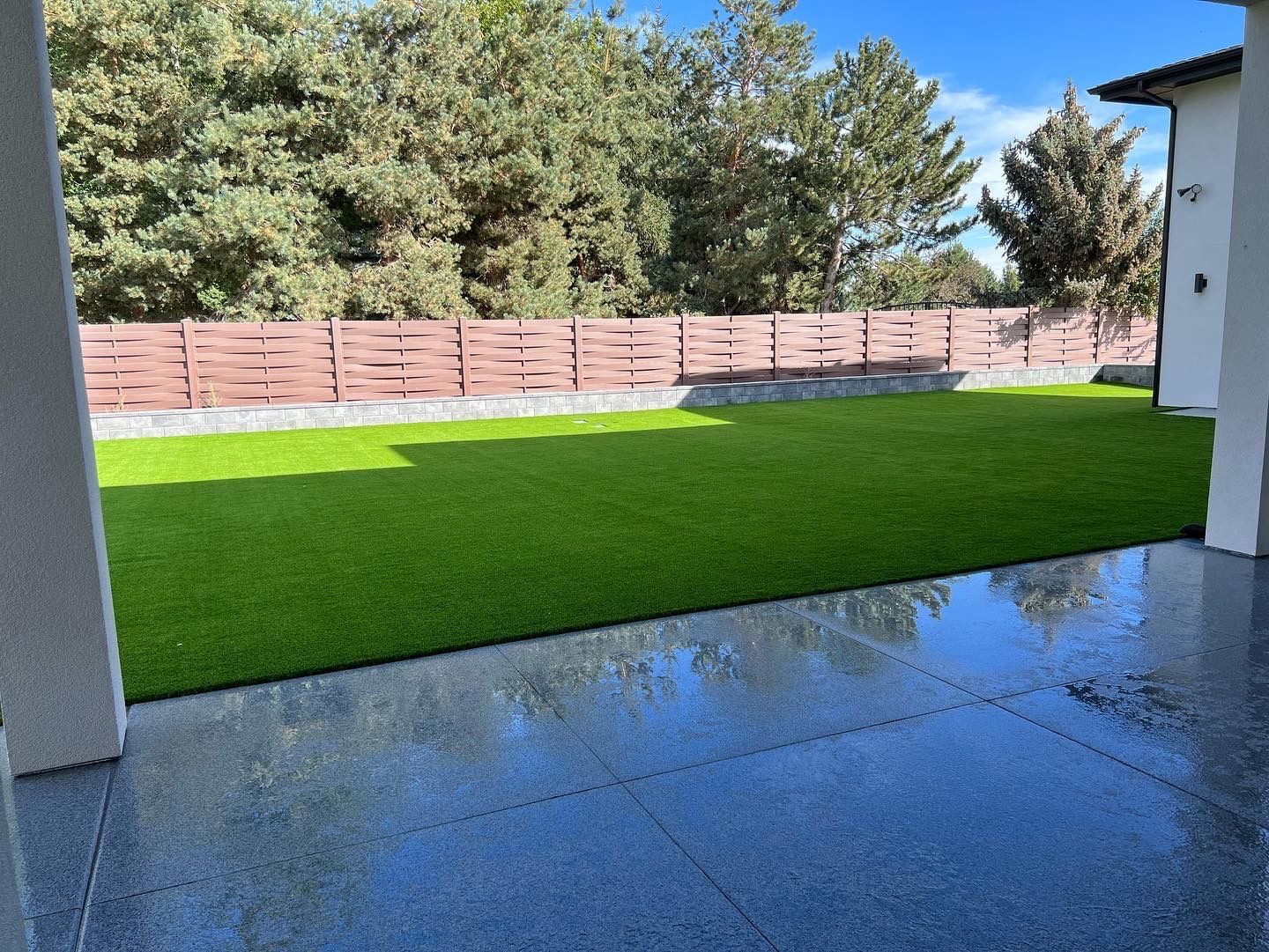 A backyard with bright green artificial grass, brown wooden fence, and a patio with a dark, speckled surface.