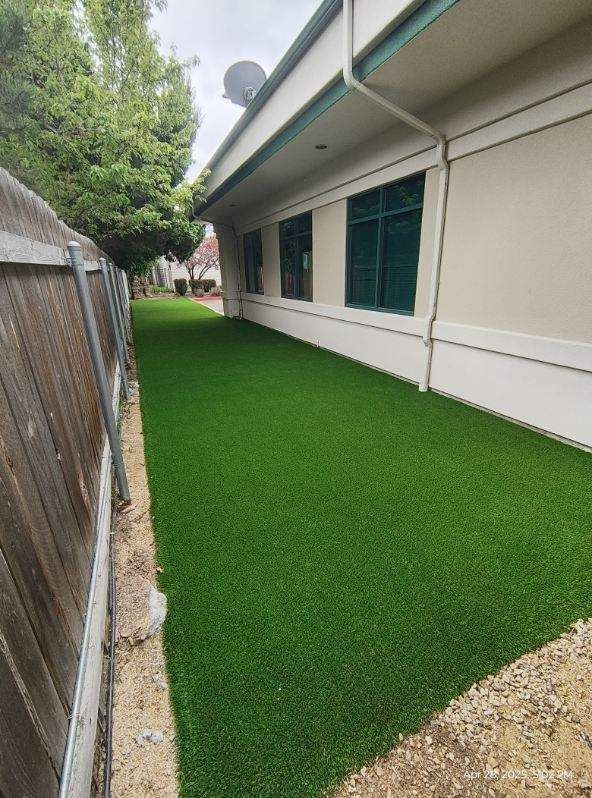 Artificial green turf along a building's side, next to a wooden fence. Overcast sky.