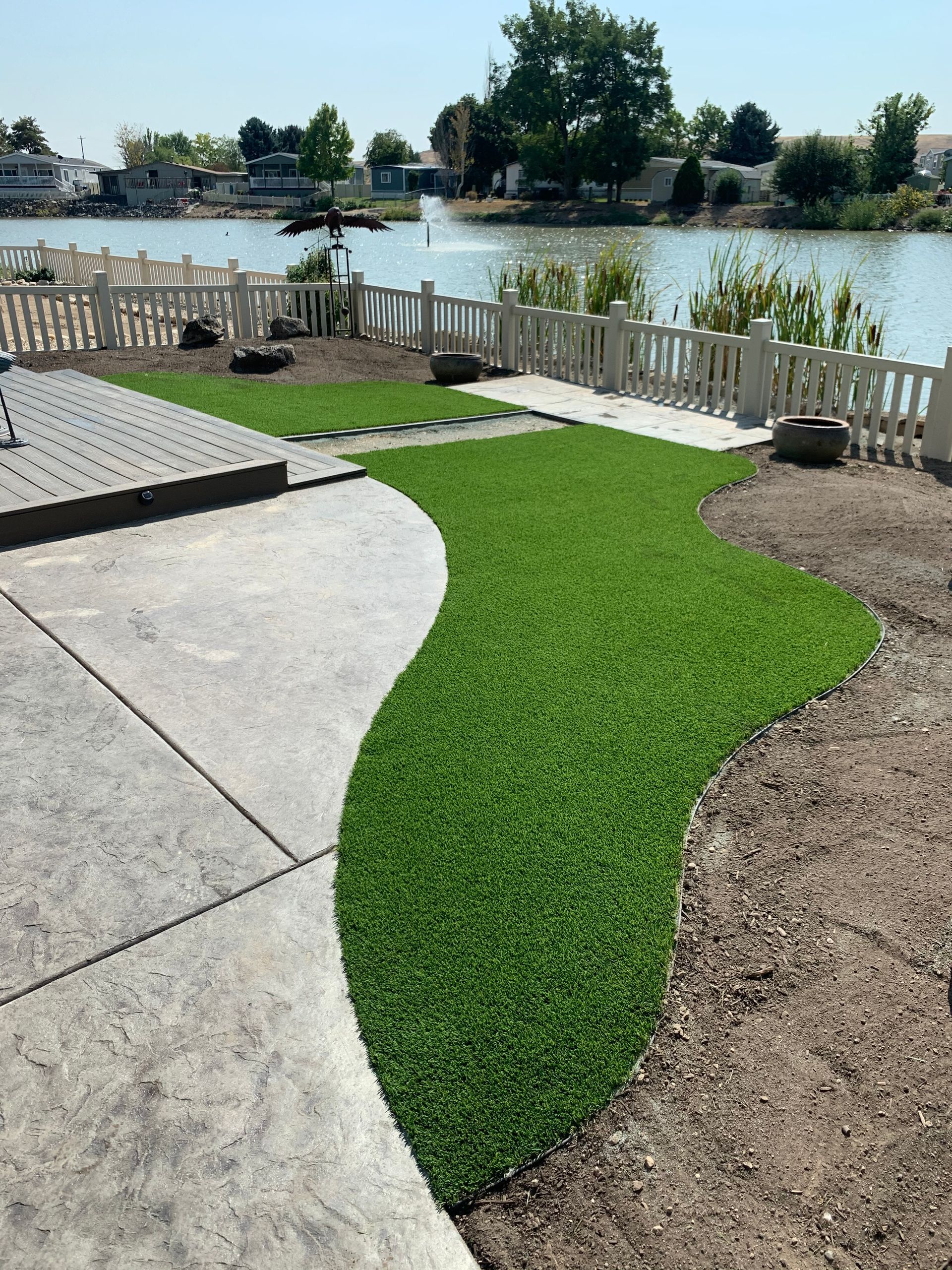 Lush green artificial turf pathway curves through a landscaped yard, bordered by concrete and a white picket fence, lake in background.