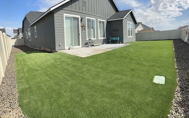 Backyard with green artificial turf, a gray house, concrete patio, and white fence.