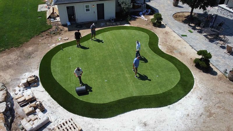 Golfers on a green artificial putting surface, surrounded by darker green and tan construction.