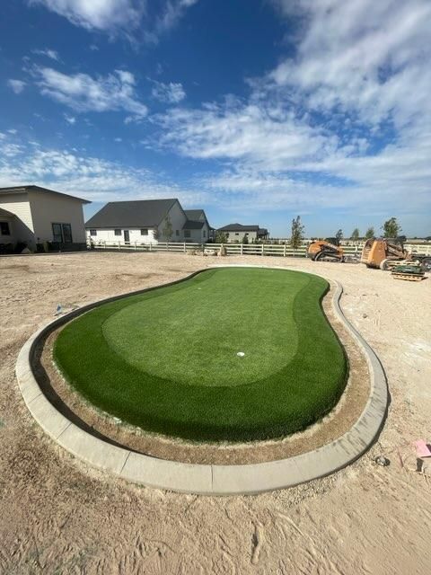 Green golf putting green with concrete border in a residential yard under a blue sky.