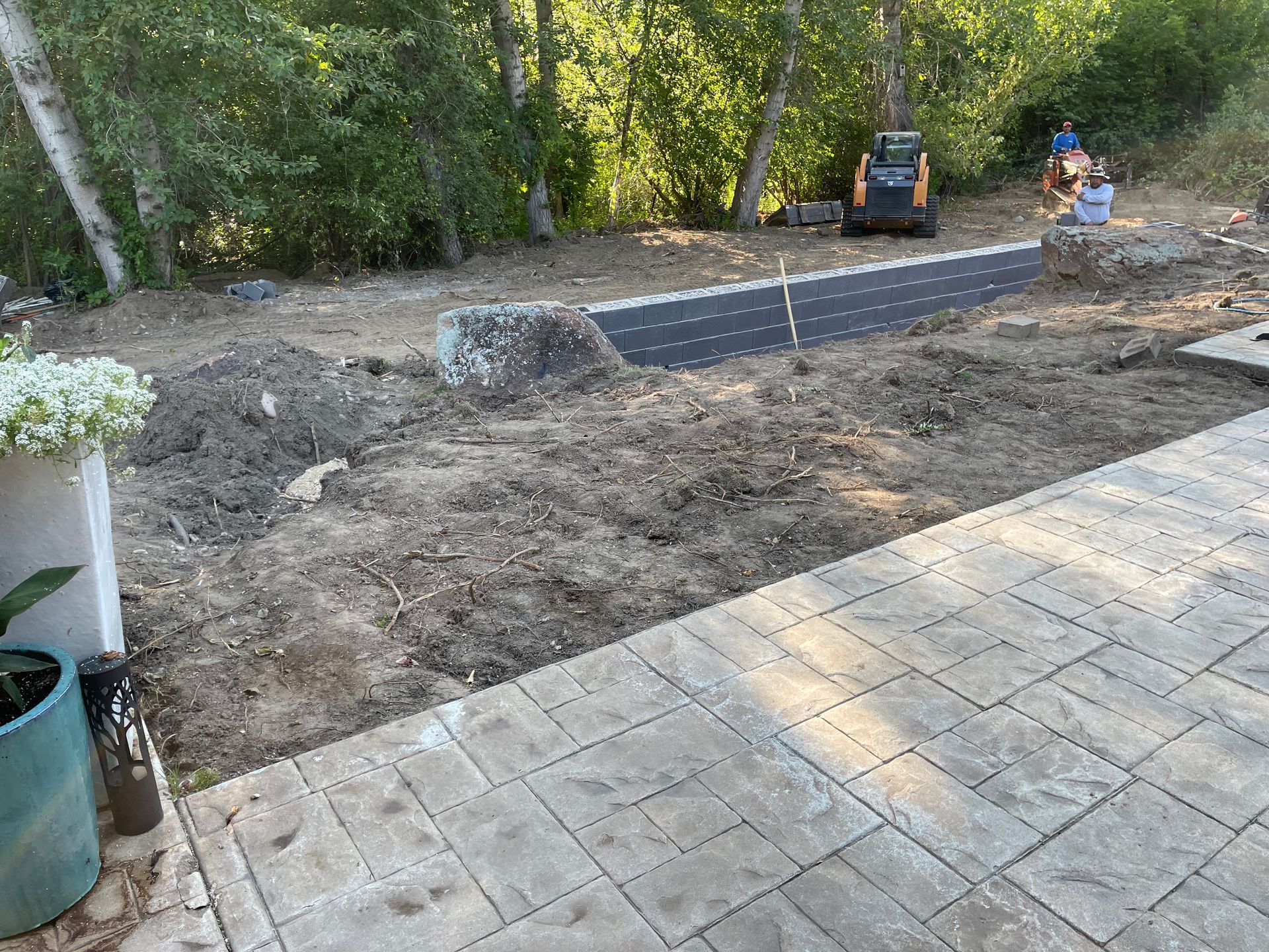 Backyard with paver patio, exposed dirt, retaining wall under construction, trees in background.