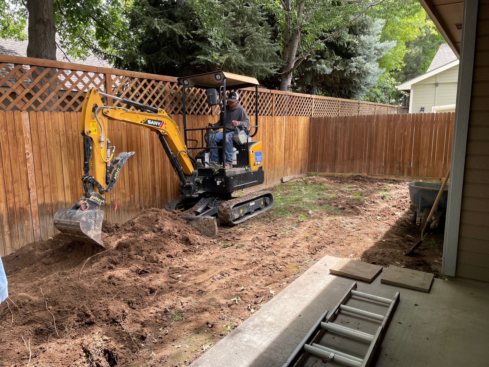 A person operating a small excavator in a backyard, digging up dirt. Wooden fence surrounds the yard.