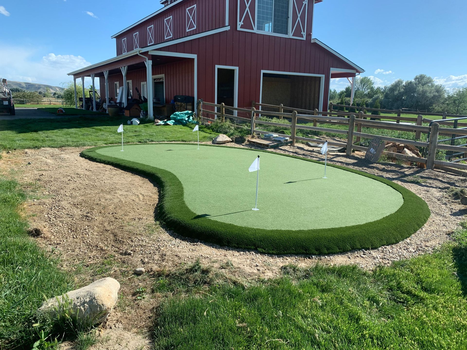 Artificial green with three flags in front of a red barn and wooden fence.
