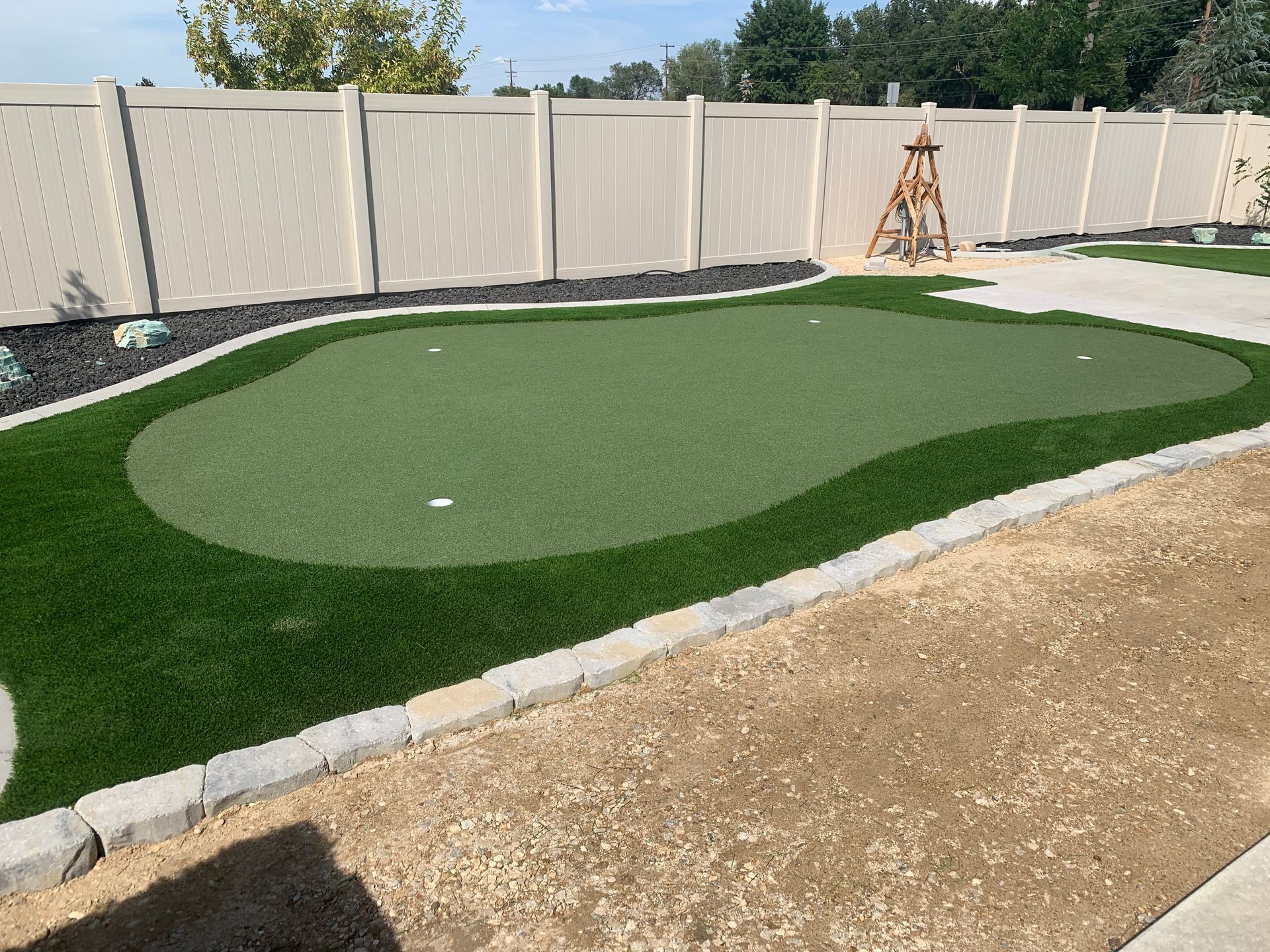 A backyard putting green with beige fence, dark border, and light gravel area.