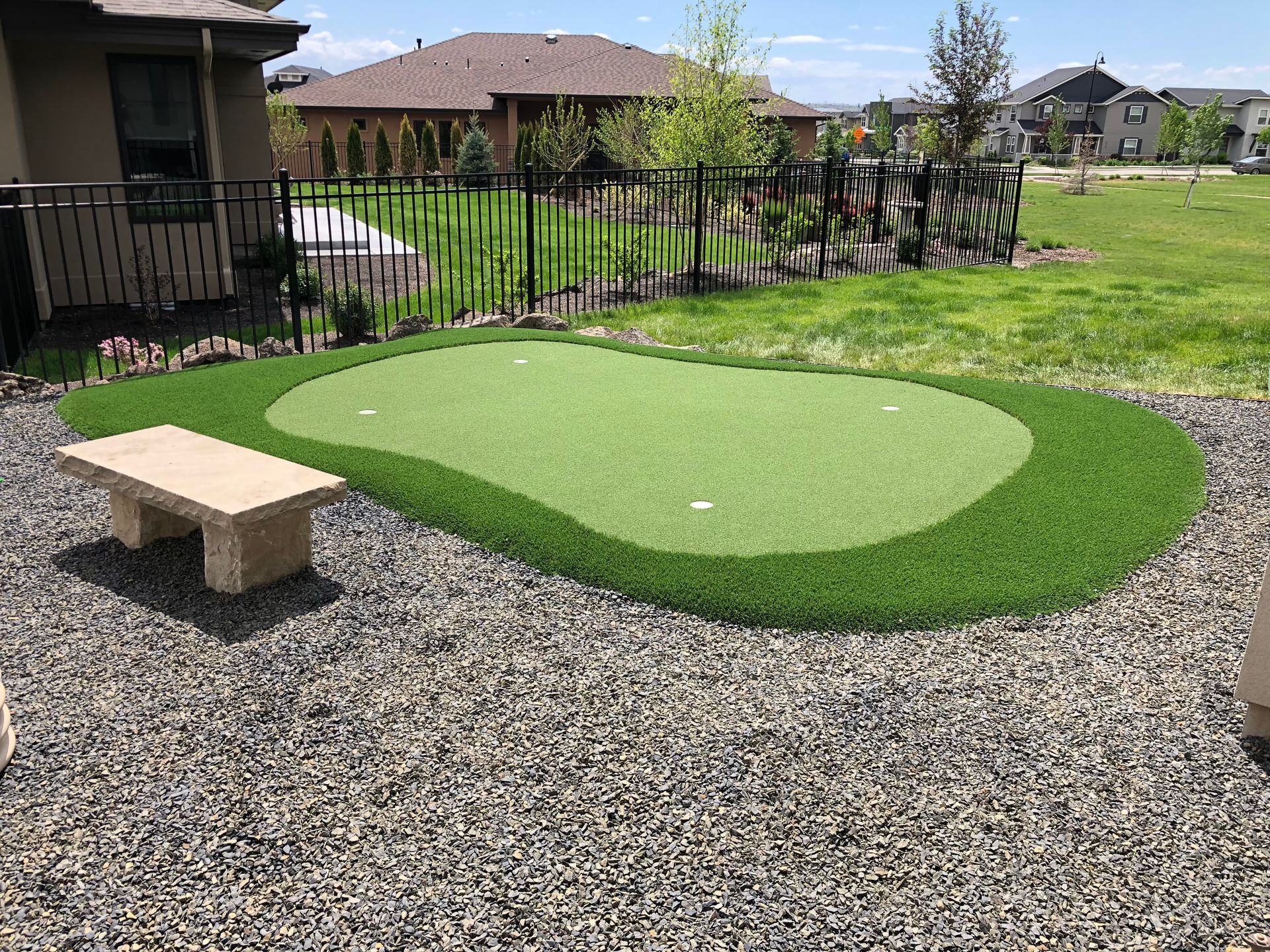 Artificial turf putting green in a backyard with stone bench, surrounded by gravel and a fence.