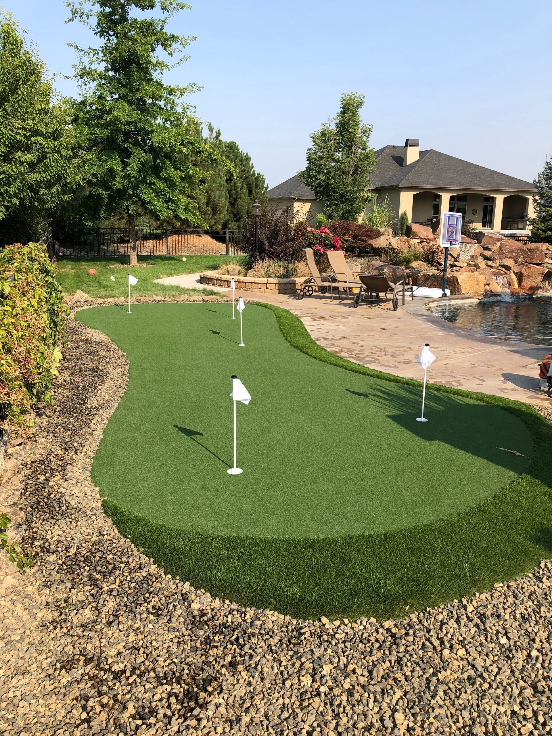 Green putting green with multiple holes and flags in a backyard setting near a house and trees.