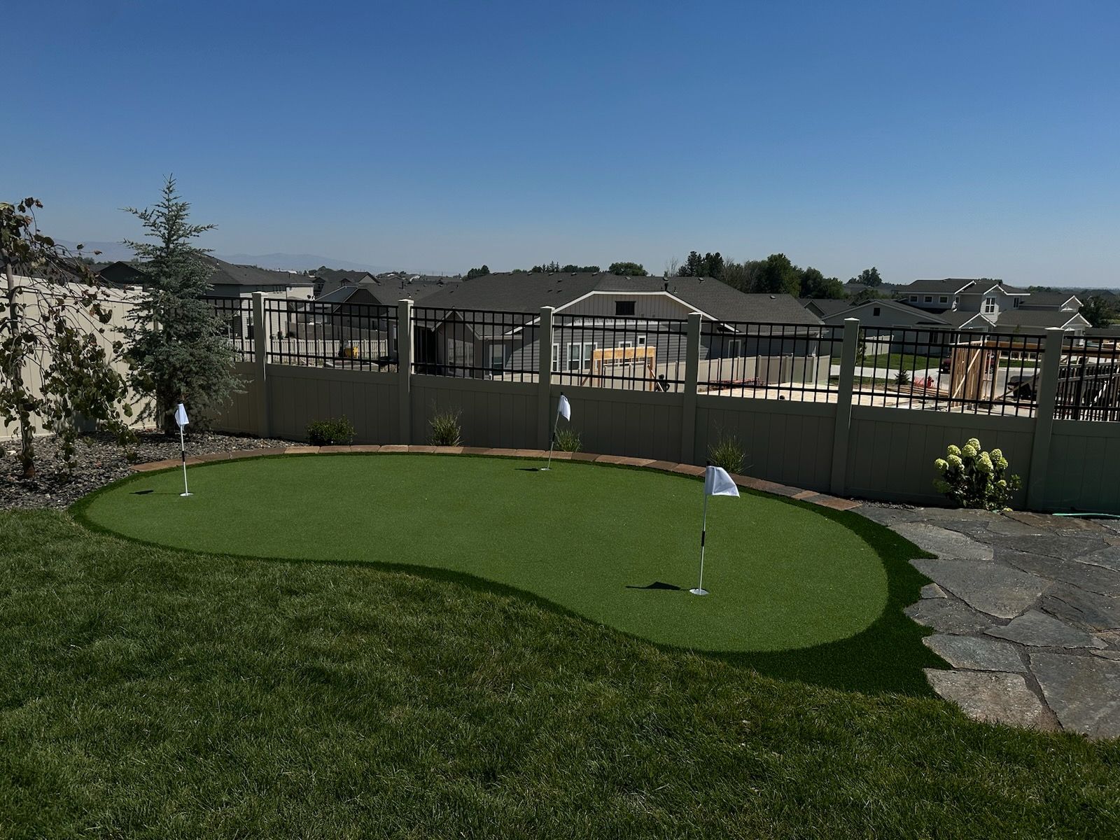 Backyard putting green with flags, surrounded by grass, trees, and a fence under a blue sky.