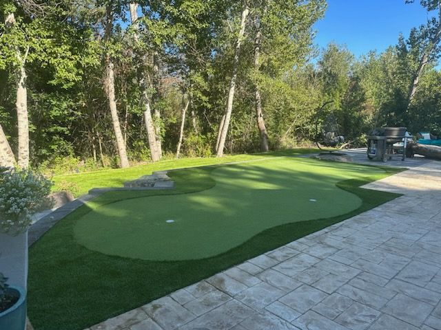 Backyard putting green, bright green turf, two holes, surrounded by trees and patio stones.