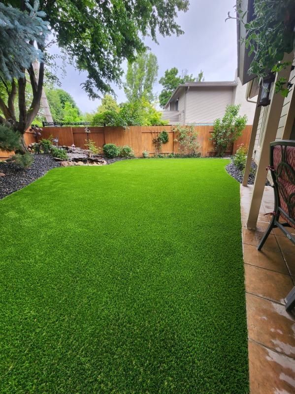 Lush green artificial lawn in a backyard. Wooden fence, trees, and small waterfall in the background.