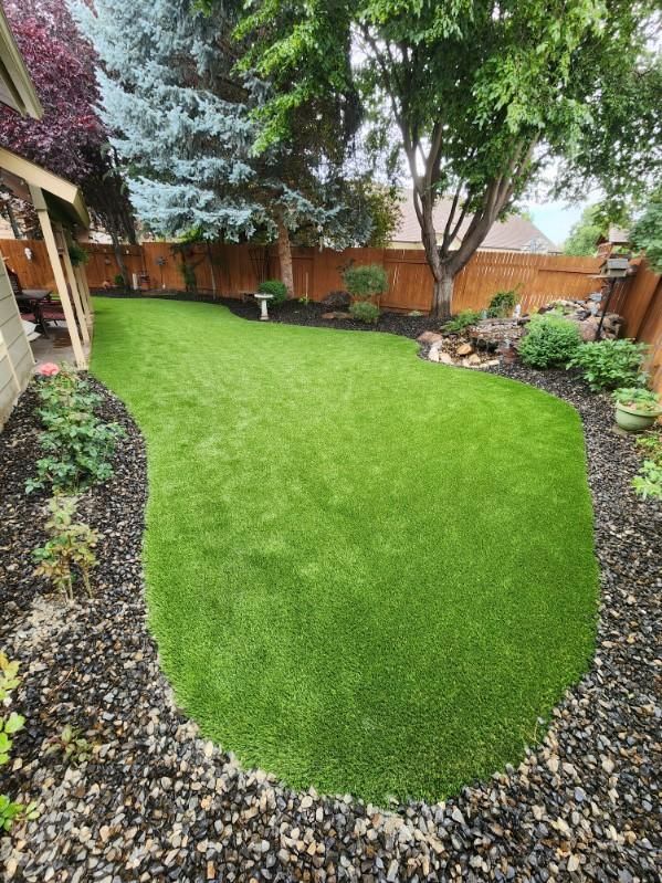 Green artificial turf lawn in a backyard, surrounded by landscaping and a wooden fence.