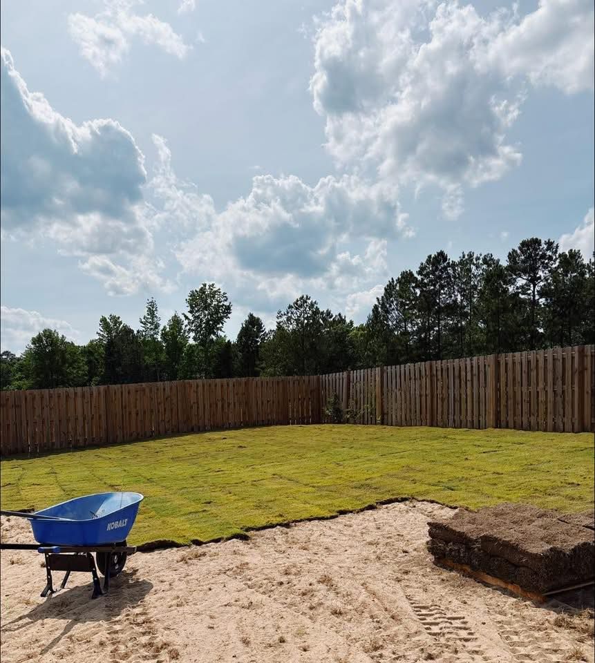 Backyard with new sod installation. Wheelbarrow, blue sky with clouds, wooden fence, trees in background.