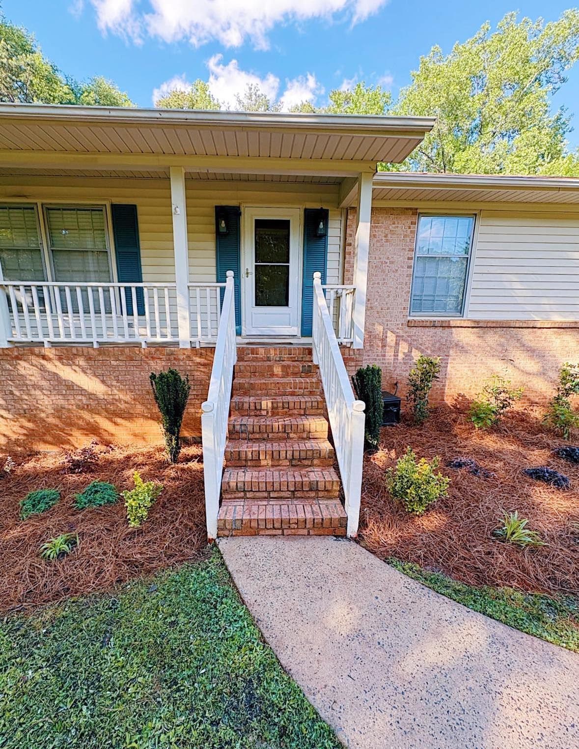 Brick steps lead to a light-colored house with a porch and blue shutters, under a partly cloudy sky.